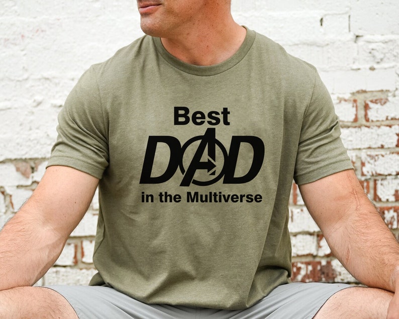 Puede incluir: Una camiseta verde con texto negro que dice "Best Dad in the Multiverse". Las letras "D" y "A" de "Dad" est&aacute;n estilizadas para parecerse al logotipo de los Avengers.