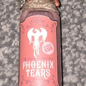 May include: A small glass bottle filled with a shimmering pink liquid, labeled "Phoenix Tears." The label features a white phoenix illustration and the words "Extremely Rare." The bottle is sealed with a cork and twine.