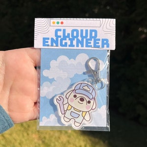 May include: A white and blue cartoon dog wearing a blue hat and overalls holding a wrench. The dog is on a clear acrylic keychain with a silver metal clasp. The keychain is packaged in a clear plastic bag with a white card that says "Cloud Engineer".
