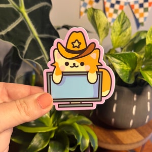 May include: A cartoon sticker of a cat wearing a cowboy hat and peeking over a television screen. The cat is orange and white with a pink nose and a pink outline. The television screen is blue and has a pink outline. The sticker is on a pink background.
