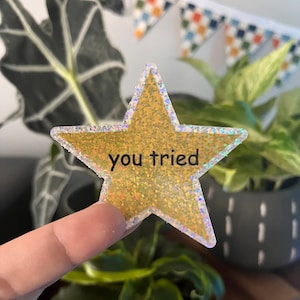 Könnte beinhalten: Ein goldener Glitzer-Stern-Aufkleber mit holografischem Rand. Der Aufkleber sagt "you tried" in schwarzem Text.