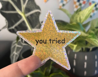 May include: A gold glitter star sticker with a holographic border. The sticker says "you tried" in black text.