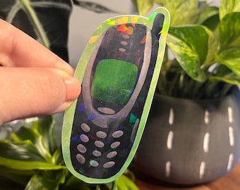 May include: A holographic sticker of a vintage cell phone with a green screen. The sticker is on a green plant.