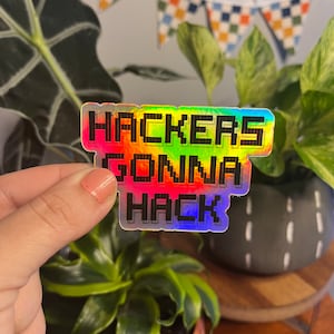 May include: A holographic sticker with the text "Hackers Gonna Hack" in a pixelated font. The sticker is rainbow colored and has a black outline.