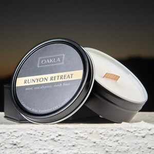 May include: A black tin candle with a white label that reads "OAKLA RUNYON RETREAT mint, eucalyptus, fresh lime Coconut Wax Essential Oil Candle". The candle is sitting on a white surface.