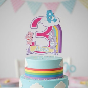 May include: A two-tiered birthday cake with light blue frosting, white cloud decorations, and a rainbow stripe. The cake is topped with a number 3 and Care Bear characters. The name "Alina" is displayed on the cake topper.