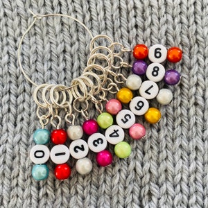 May include: A set of ten colourful stitch markers with numbers 0-9 on white beads. The markers are attached to a silver ring.