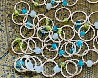 May include: A close-up of a group of silver rings with colorful beads. The beads are blue, green, and light blue. The rings are arranged in a random pattern.