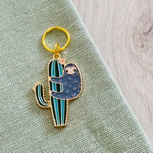 May include: A gold-tone keychain featuring a cartoon sloth hanging on a blue and green cactus. The keychain has a gold ring for attaching to keys or a bag.