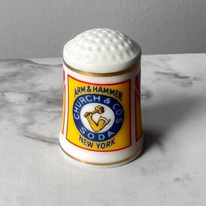 May include: A white ceramic thimble with a dimpled top and a gold rim. The thimble features the "ARM & HAMMER CHURCH & CO SODA NEW YORK" logo in a yellow and blue design with red accents.
