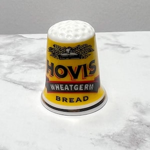 May include: A yellow ceramic thimble with a white top. The thimble has a black and red logo that says "Hovis Wheatgerm Bread".