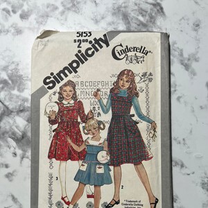 May include: Simplicity sewing pattern number 5153 for a girl's dress. The pattern includes three different dress styles: a red dress with white polka dots, a blue dress with white trim, and a plaid dress. The pattern is for sizes 6-child. The pattern is titled "Cinderella".