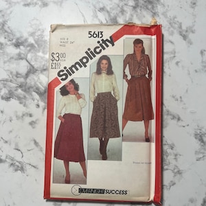 Puede incluir: Patrón de costura Simplicity número 5613 para una falda. El patrón es para una talla 8, cintura 61 cm, y es para mujeres. El patrón muestra tres estilos diferentes de faldas, todas con cintura alta. El patrón es rojo y blanco con el texto "Simplicity" en letras blancas grandes.
