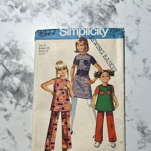 May include: Vintage Simplicity sewing pattern number 9477 for a child's sewing basket. The pattern includes three different outfits: a red and black patterned pantsuit, a green dress with red pants, and a striped dress with a black belt.