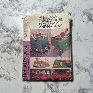 May include: A vintage McCall's sewing pattern for all-season tablecloths, placemats, and napkins. The pattern includes five sets of interchangeable trims, plus a Christmas tree skirt. The pattern cover features a collage of images showing different table settings and close-ups of the designs.