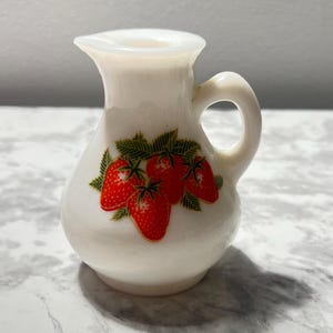 May include: A small, white ceramic pitcher with a handle and a spout. The pitcher is decorated with a cluster of red strawberries and green leaves. The pitcher is likely used for serving cream or syrup.