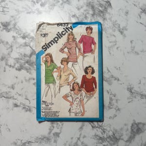 Puede incluir: Patrón de costura Simplicity vintage con ilustraciones de tops. El sobre es blanquecino con un borde azul y el logotipo de Simplicity. El número de patrón es 6432. Las ilustraciones muestran tops en rosa, verde, beige y telas estampadas.