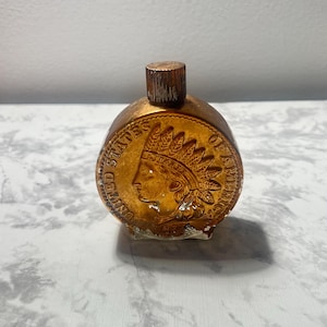 May include: A small, round, amber-colored glass bottle with a wooden stopper. The bottle features a detailed embossed design of an Indian head coin. The words "UNITED STATES OF AMERICA" are visible around the coin's edge.