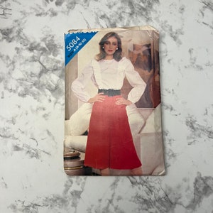 May include: Vintage sewing pattern for a blouse and skirt. The pattern is for size A (8-10-12) and is labeled 5084. The model is wearing a white blouse with a ruffle collar and a red skirt with a wide black belt.