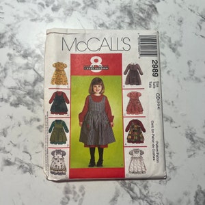 May include: McCall's sewing pattern for a girl's dress, pattern number 2989, size CD (2-3-4). The pattern includes eight different styles of dresses, including a plaid dress with a pinafore overlay, a floral print dress, and a solid color dress with a ruffle trim.