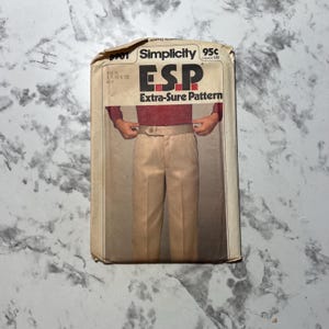 May include: A vintage Simplicity sewing pattern packet featuring a man wearing tan pants and a burgundy shirt. The packet has the text "ESP Extra-Sure Pattern" and the price "95¢". The packet is beige and has a worn appearance.