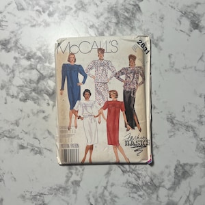 May include: Vintage McCall's sewing pattern featuring illustrations of women's fashion designs. The pattern packet shows various dress styles, including a long-sleeved blue dress, a polka-dot ensemble, and a red dress. The text "Fashion Basics" is visible.