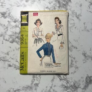 May include: Vintage McCall's printed pattern for a Misses' blouse set. The pattern features illustrations of three blouse designs. The packaging is off-white with a green and yellow stripe on the left side. The text "NEW Sizing" is in a pink box.