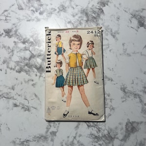 May include: Vintage sewing pattern for a girl's skirt and blouse. The pattern is from Butterick and is number 2412. The pattern is for a size 6, with a 24 inch bust. The pattern shows a girl wearing a yellow vest, a plaid skirt, and a white blouse.