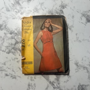 May include: A vintage sewing pattern featuring a woman in an orange dress. The pattern is from the "New York Designers' Collection Plus" and is labeled "N1056" with the designer's name "Geoffrey Beene". The pattern's size is 14, bust 36.