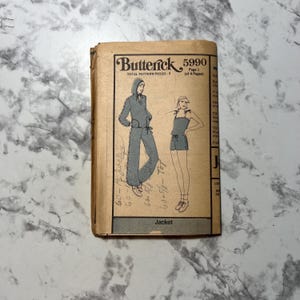 May include: A vintage Butterick sewing pattern, number 5990, featuring illustrations of a hooded jacket and a romper. The cover is tan with black text and drawings. The pattern includes measurements and is for a jacket.