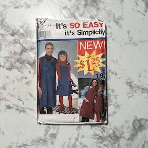 May include: A sewing pattern package featuring a woman and a girl wearing denim jumpers over red turtlenecks. The package has the text "It's SO EASY it's Simplicity" and "NEW!" in a red starburst.
