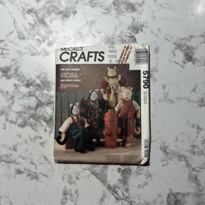 May include: A McCall's Crafts sewing pattern for making cat-themed stuffed toys. The pattern is called "The Cat's Meow" and features a variety of cat designs, including a calico cat, a tabby cat, and a tuxedo cat. The pattern is printed on a white background with red and blue text.