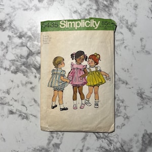 Puede incluir: Patrón de costura vintage Simplicity 5435 para un vestido de niña pequeña. El patrón es para las tallas 14, 19 y 19. El patrón muestra a tres niñas usando diferentes versiones del vestido. El vestido es un estilo simple y sin mangas con una falda fruncida. El patrón está impreso en un fondo blanco con texto verde y negro.
