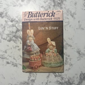 May include: A Butterick 6029 sewing pattern envelope titled Luv 'n Stuff featuring illustrations of two fabric rabbit dolls with long ears, wearing ruffled bonnets and dresses, with one doll standing and one doll sitting in a pink toy cart.