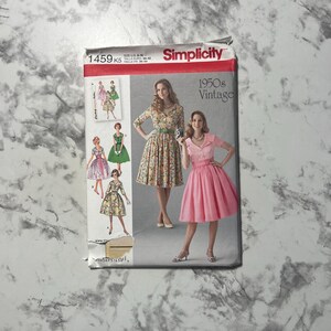 May include: A vintage Simplicity sewing pattern for 1950s dresses. The pattern features illustrations of women in various dress styles, with a woman in a floral dress and a woman in a pink dress on the front. The pattern size is US 8-16.