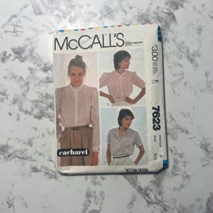 May include: A McCall's 7623 sewing pattern envelope featuring three variations of a blouse. The cover displays illustrations of a long-sleeved blouse with a ruffled collar, a short-sleeved blouse with a rounded collar, and a short-sleeved collared shirt.