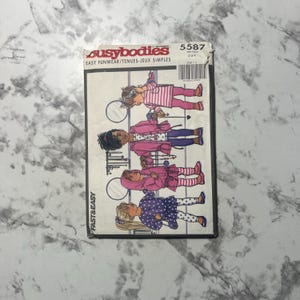 May include: A vintage sewing pattern by "busybodies" featuring illustrations of children's clothing. The pattern shows drawings of children in coats, dresses, and leggings. The cover is a mix of pink, purple, and white.