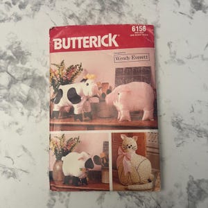 May include: A Butterick 6156 sewing pattern envelope designed by Wendy Everett. The front features illustrations of four stuffed animal toys: a black and white cow, a pink pig, a white sheep with black legs, and a floral print cat with a pink bow.