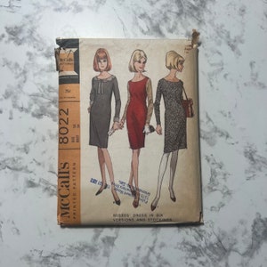 May include: Vintage McCall's printed sewing pattern, size 14, featuring illustrations of three dresses. The pattern cover is tan with the McCall's logo and text. The dresses are in shades of gray, red, and black. The text reads "Misses' Dress in Six Versions and Stockings."