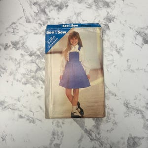 May include: A vintage See & Sew pattern for a girl's dress. The pattern number is 3286, size A (2-3-4). The dress is blue with white straps and a white ruffle trim. The model is wearing a white blouse with a white collar.
