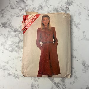 Könnte beinhalten: Vintage Schnittmuster mit einer Frau in einem langärmeligen Kleid mit Gürtel. Die Schnittmusterhülle ist beige mit rotem Text "STITCH 'N SAVE". Das Kleid ist rotbraun mit einem kleinen Tupfenmuster.