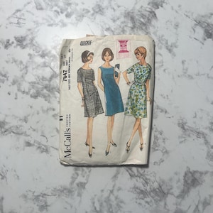 Könnte beinhalten: Vintage McCall's Schnittmuster mit Illustrationen von drei Kleidern. Die Schnittmusterpackung ist cremefarben mit Text und Illustrationen in Schwarz, Rosa und Blau. Die Schnittmusternummer ist 7647, und die Größe ist 14W, Brustumfang 89 cm.