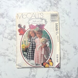 May include: McCall's sewing pattern for a doll dress, size CD (2, 3, 4). The pattern is titled "Ruffles and Lace Treasured Collection" and features a pink and white gingham dress and a black and white gingham dress.