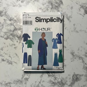May include: A Simplicity sewing pattern packet featuring a woman in a blue outfit. The packet includes illustrations of various clothing designs, including dresses, jackets, and pants. The text "Simplicity" and "2 HOUR" are prominently displayed on the front.