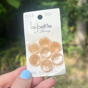 May include: A card of seven round, light peach-colored buttons. The card reads "la petite by Lansing" and includes the size and number of buttons. The buttons have a pearlescent sheen and are arranged in a circular pattern.