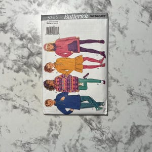 May include: A Butterick sewing pattern with illustrations of children's clothing designs. The cover shows a pink hooded sweatshirt, an orange dress, a patterned tunic, and a blue jacket with striped trousers. The pattern number is 3715.