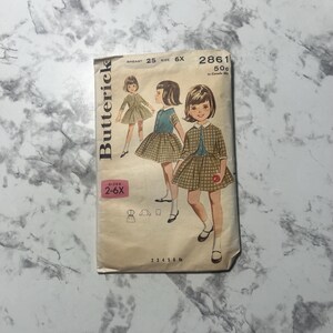 May include: Vintage sewing pattern for a girl's dress and jacket. The pattern is for sizes 2-6X and includes instructions for making a plaid skirt and a matching jacket. The pattern is from Butterick and has the number 2861 on it.