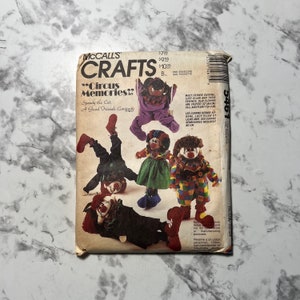 Könnte beinhalten: Nähmuster von McCall's Crafts für die Herstellung von vier Zirkus-Clown-Puppen. Das Muster trägt den Titel "Circus Memories" und zeigt eine Katze namens Speedy. Das Muster enthält Anleitungen für die Herstellung einer lila, roten, grünen und schwarzen Clownpuppe.