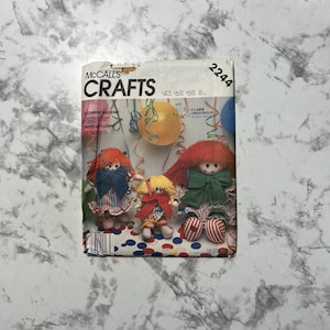Peut inclure: Un patron de couture vintage McCall's Crafts pour fabriquer des poupées clowns. La couverture du patron présente trois poupées clowns faites à la main avec des cheveux rouges, jaunes et verts, et des tenues colorées. Le texte sur la couverture indique "CLOWNIN' AROUND" et "FAITES LE CLOWN".