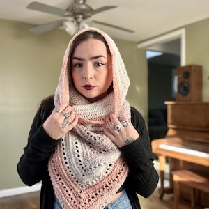 May include: A hooded scarf in shades of pink, gray, and white, crocheted with a delicate stitch pattern. The scarf is worn by a person with dark hair and a black shirt.